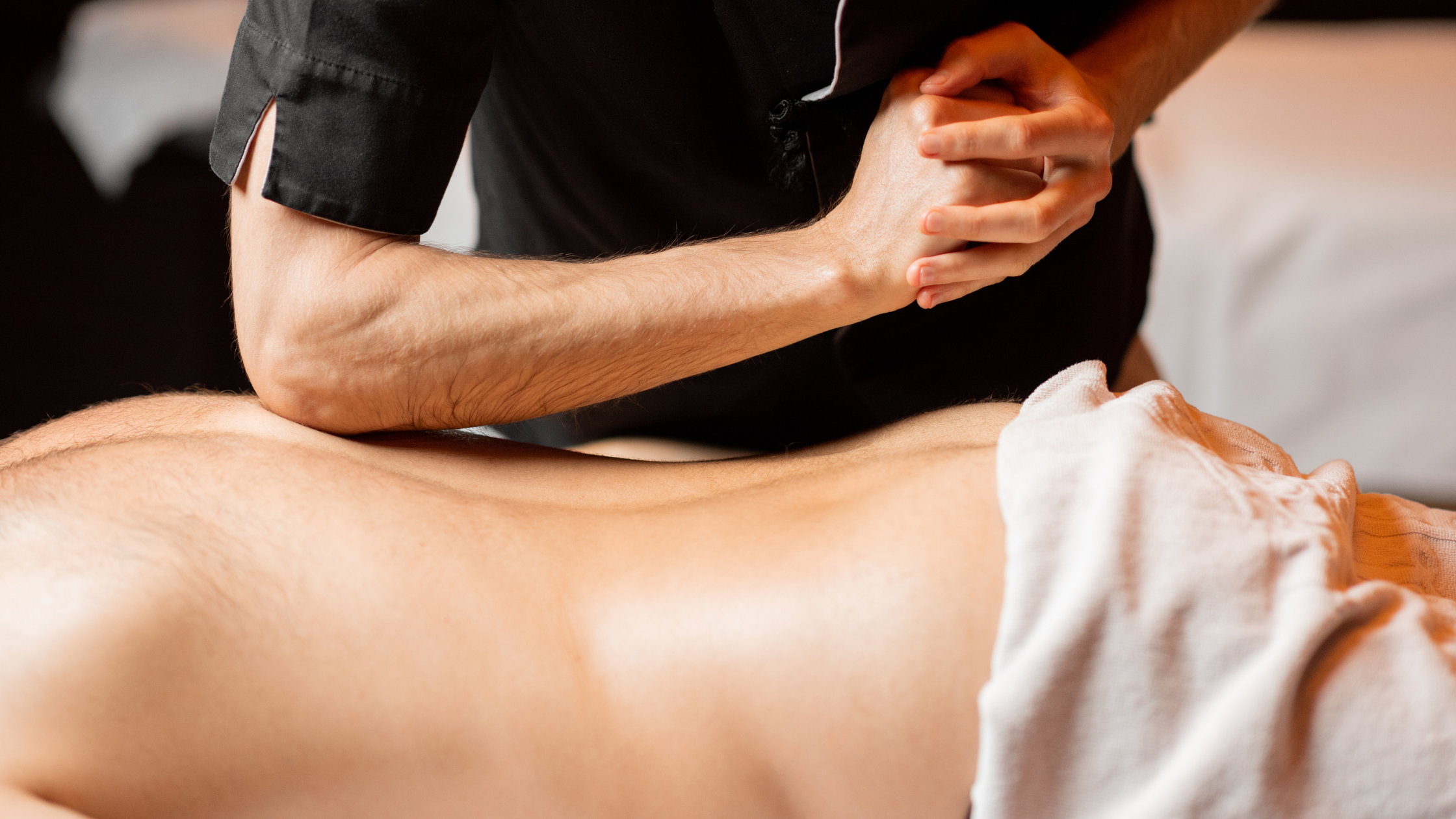 A person lying on a massage table, receiving a deep tissue massage on their back. The massage therapist is using their elbow to apply firm pressure to the muscles.